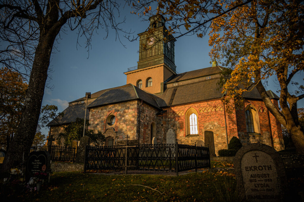 n storslagen bild av Vårfrukyrkan i Enköping, fångad i gyllene eftermiddagsljus. Kyrkans majestätiska arkitektur kontrasteras av de omgivande höstträdens varma färger och den historiska kyrkogården som viloplats.