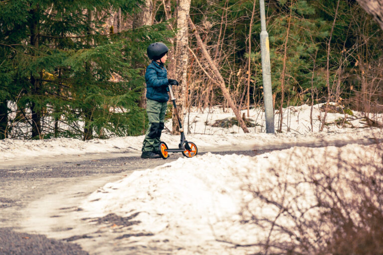 En liten pojke klädd i en mörkgrön jacka och svarta byxor står redo på sin orange och svarta sparkcykel. Bakom honom vilar en smältande snödriva vid kanten av en skogsväg, signalerande tidig vår i Enköping.
