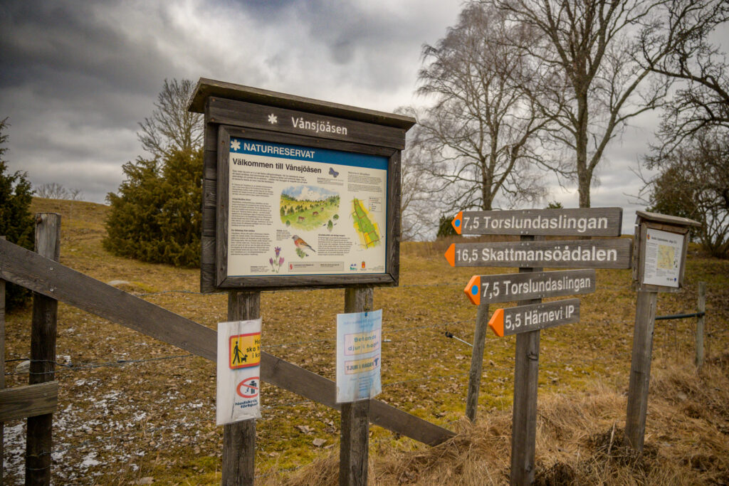 Informationskylt vid naturreservatet Vånsjöbroåsen som visar vägskyltar och karta, omgiven av barträd och molntäckt himmel.