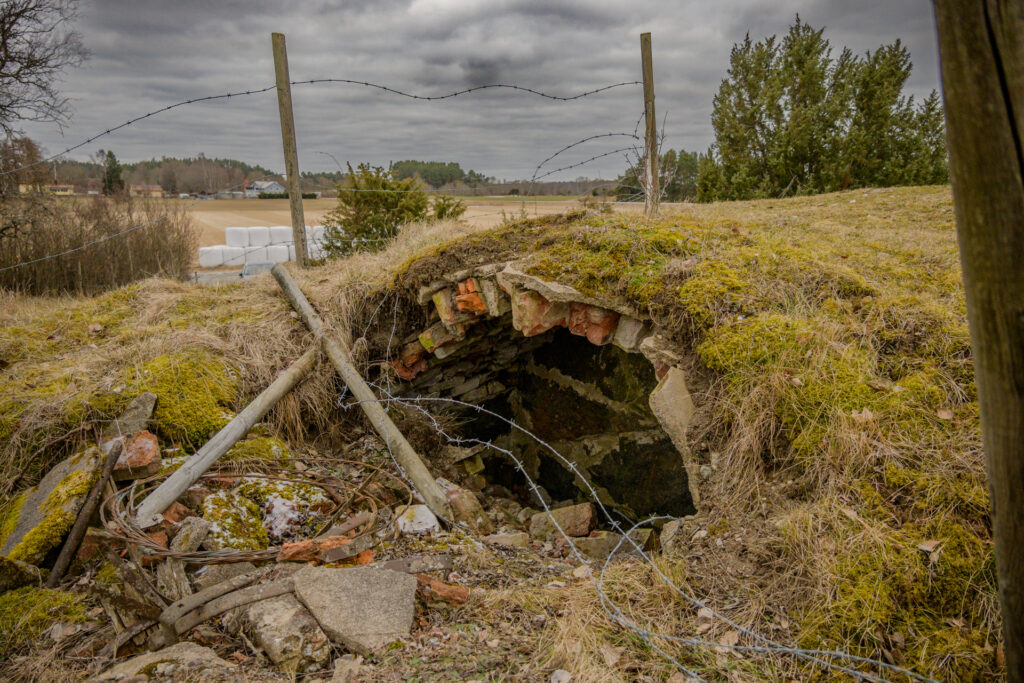 Gamla jordkällaren i Vånsjöbroåsen omgivna av vildgräs och taggtråd, vilket indikerar en rik historia i området nära Enköping.