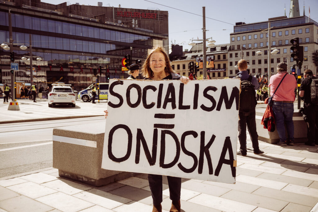 Lena Malmberg håller upp en skylt med texten "SOCIALISM = ONDSKA" vid en demonstration arrangerad av Medborgerlig Samling på en solig dag i en stadsmiljö, symboliserande engagemang för individuell frihet och motstånd mot socialism i Sverige.
